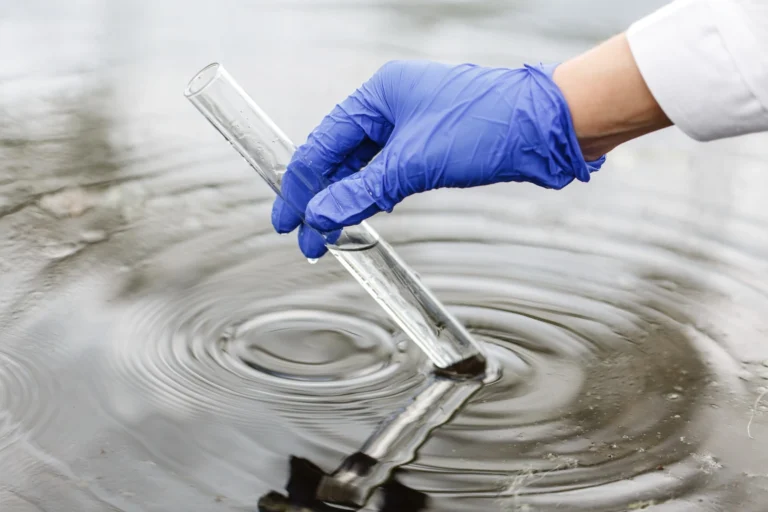 Hand with blue glove collecting water sample in test tube.